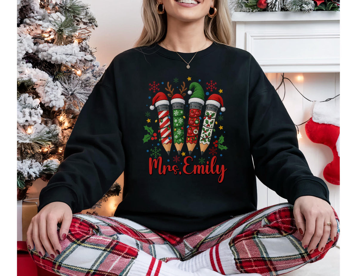 May include: Black sweatshirt with a Christmas pencil design. The pencils are decorated with festive patterns and Santa hats. The name "Mrs. Emily" is written in red script below the pencils. The person is wearing red and white plaid pajama pants.