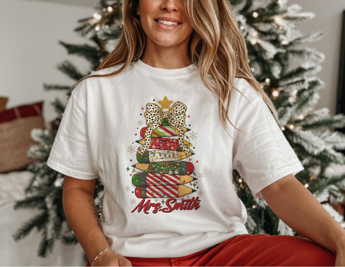May include: White t-shirt featuring a Christmas tree design made of pencils, presents, and a bow. The design includes a star, lights, and the name "Mrs. Smith" in red script. The shirt is a comfortable, casual style.