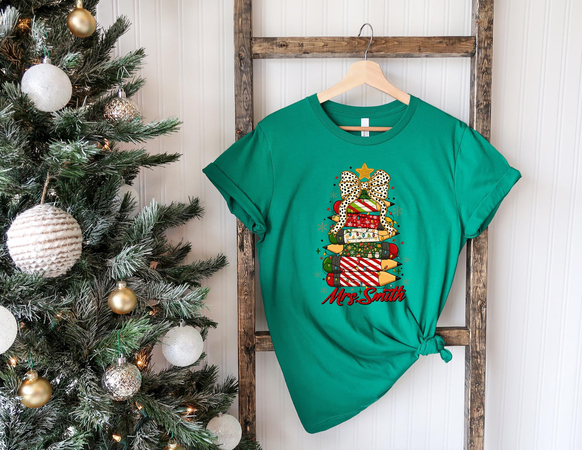 May include: A green t-shirt featuring a Christmas tree design made of pencils and a bow. The pencils are decorated with various patterns and colors. The text "Mrs. Smith" is written in red script below the tree.