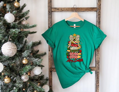 May include: A green t-shirt featuring a Christmas tree design made of pencils and a bow. The pencils are decorated with various patterns and colors. The text "Mrs. Smith" is written in red script below the tree.