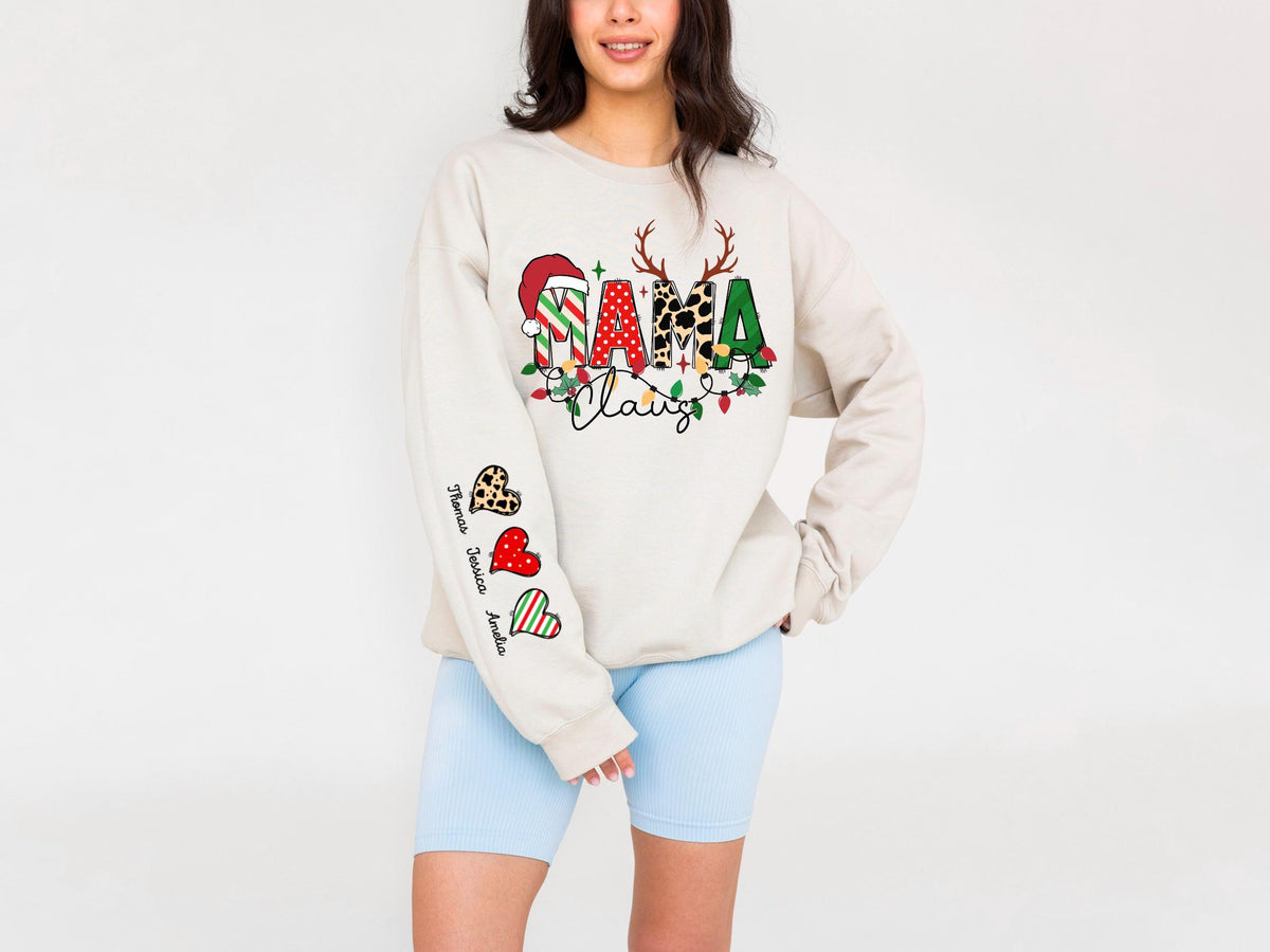 May include: Beige long-sleeve sweatshirt with a colorful "Mama Claus" design. The design includes a Santa hat, reindeer antlers, and festive lights. The left sleeve features heart-shaped graphics with names. The sweatshirt is paired with light blue shorts.