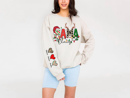 May include: Beige long-sleeve sweatshirt with a colorful "Mama Claus" design. The design includes a Santa hat, reindeer antlers, and festive lights. The left sleeve features heart-shaped graphics with names. The sweatshirt is paired with light blue shorts.
