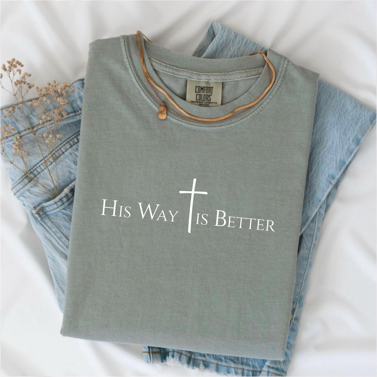 May include: Sage green Comfort Colors t-shirt with the text "His Way Is Better" and a white cross graphic. A gold-colored necklace with a small pendant rests on the shirt, which is folded on top of a pair of light blue jeans.