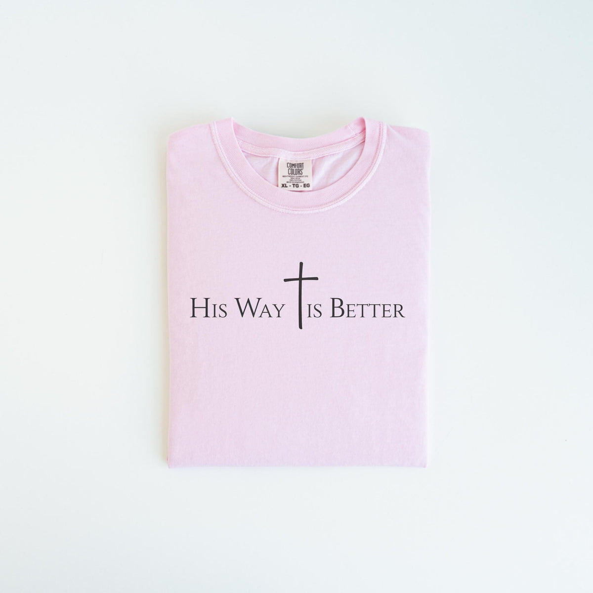 May include: A folded light pink t-shirt with the text "His Way Is Better" and a cross graphic in black. The shirt is made of a soft material and has a crew neck.