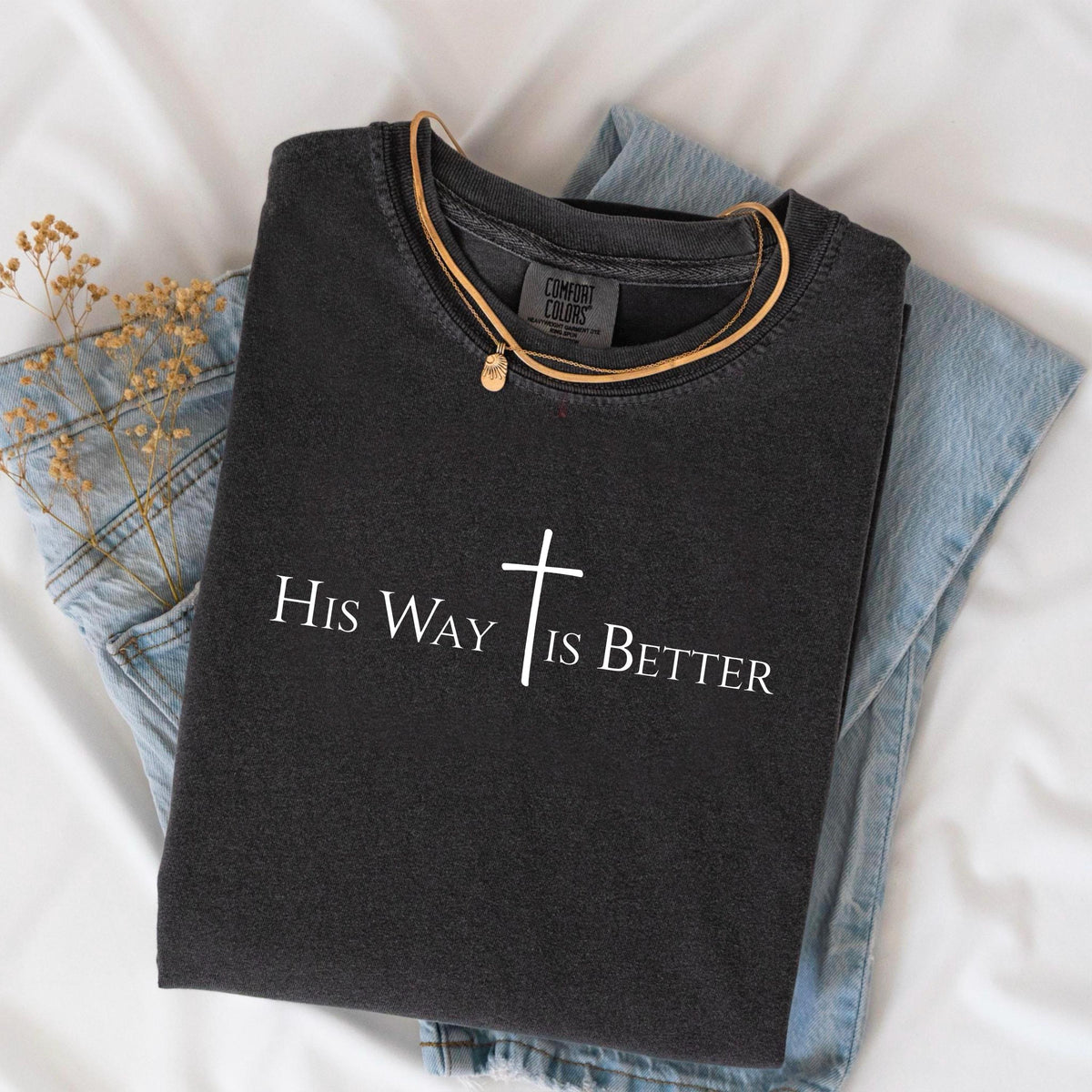 May include: Dark gray t-shirt with the text "His Way Is Better" and a cross graphic. The shirt is folded and placed on top of blue jeans. A gold necklace with a pendant is draped over the shirt.