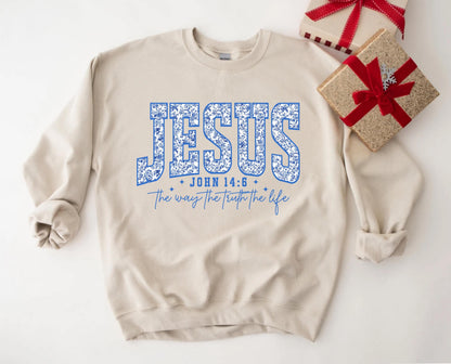May include: A beige sweatshirt with the word "JESUS" in large, blue floral letters. Below, it reads "JOHN 14:6" and "The way the truth the life" in blue script. Two wrapped gifts with red ribbons are in the background.