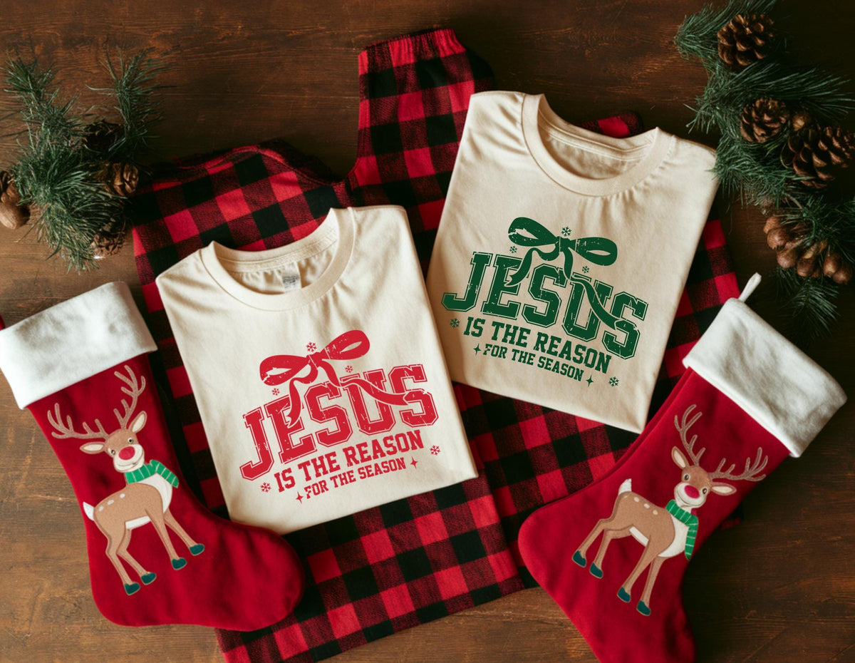 May include: Two cream-colored t-shirts with the text "JESUS IS THE REASON FOR THE SEASON" in red and green, along with a pair of red and black plaid pajama pants. Two red Christmas stockings with reindeer designs are also present.