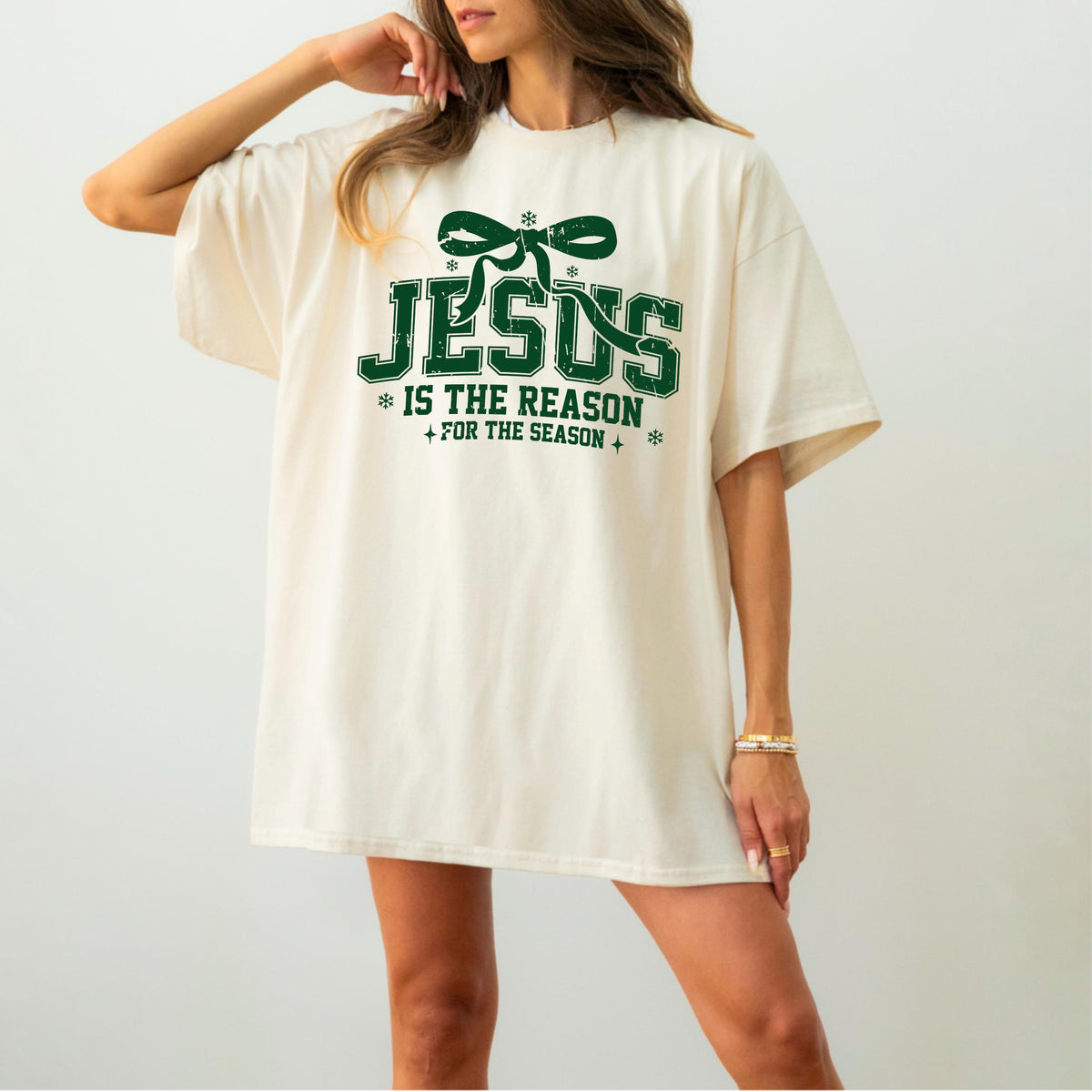 May include: Cream-colored t-shirt with the text "JESUS IS THE REASON FOR THE SEASON" in green. The design includes a green bow and snowflakes. The shirt has short sleeves and a relaxed fit.