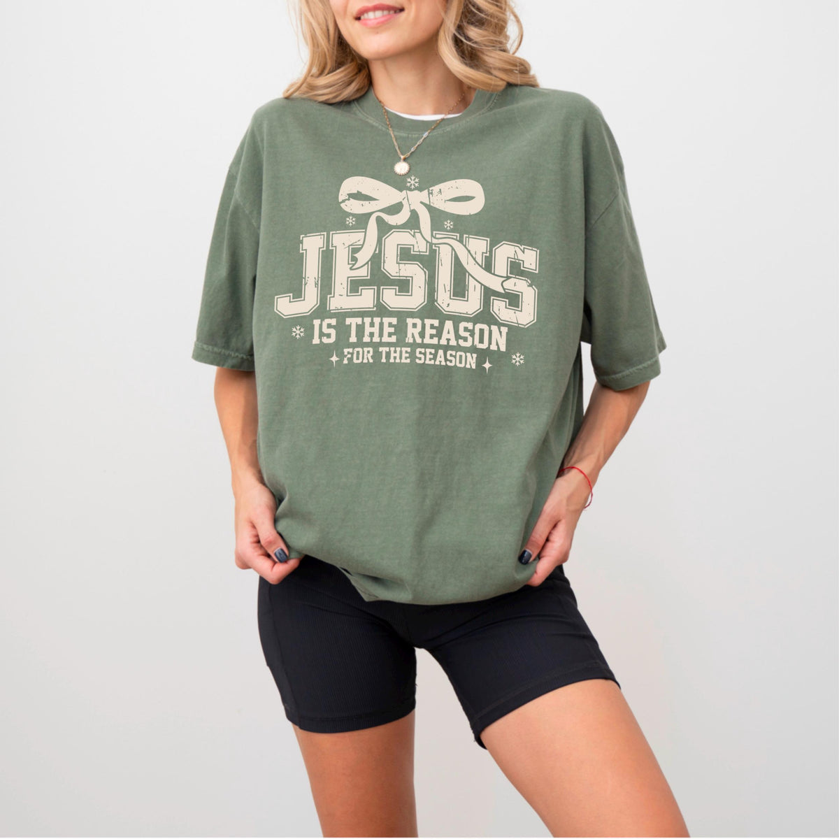 May include: Olive green t-shirt with the text "JESUS IS THE REASON FOR THE SEASON" in a cream-colored, vintage-style font. The design includes a bow and snowflakes. The shirt is paired with black shorts.