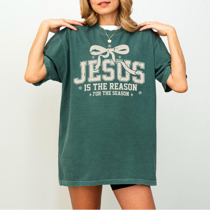 May include: A dark green t-shirt with the text "JESUS IS THE REASON FOR THE SEASON" in a distressed, off-white font. The design includes a bow and snowflakes. The shirt is a relaxed fit.