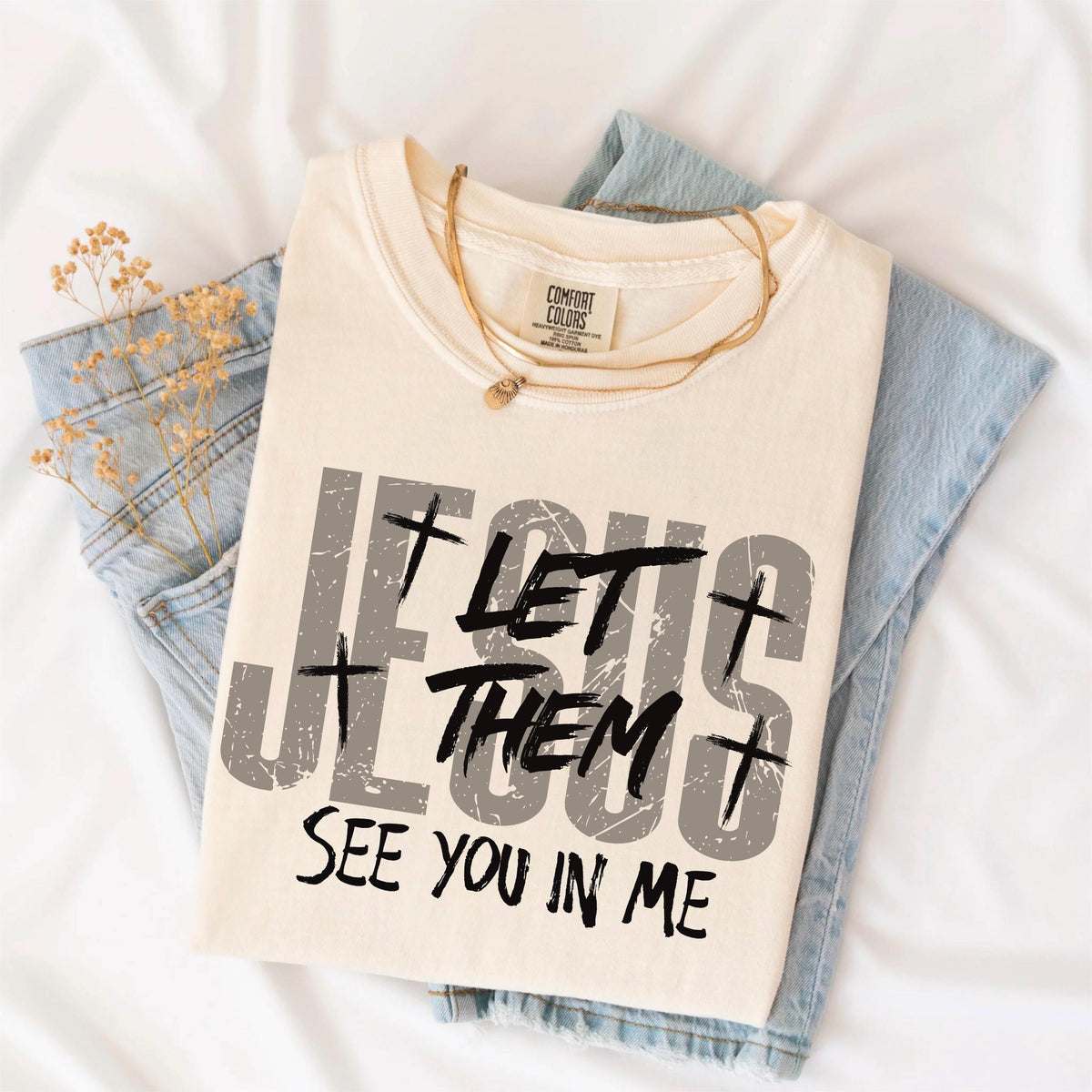 May include: Cream-colored t-shirt with the text "JESUS LET THEM SEE YOU IN ME" in distressed gray and black lettering. The shirt is folded and rests on a pair of blue jeans. A gold necklace is draped across the shirt.