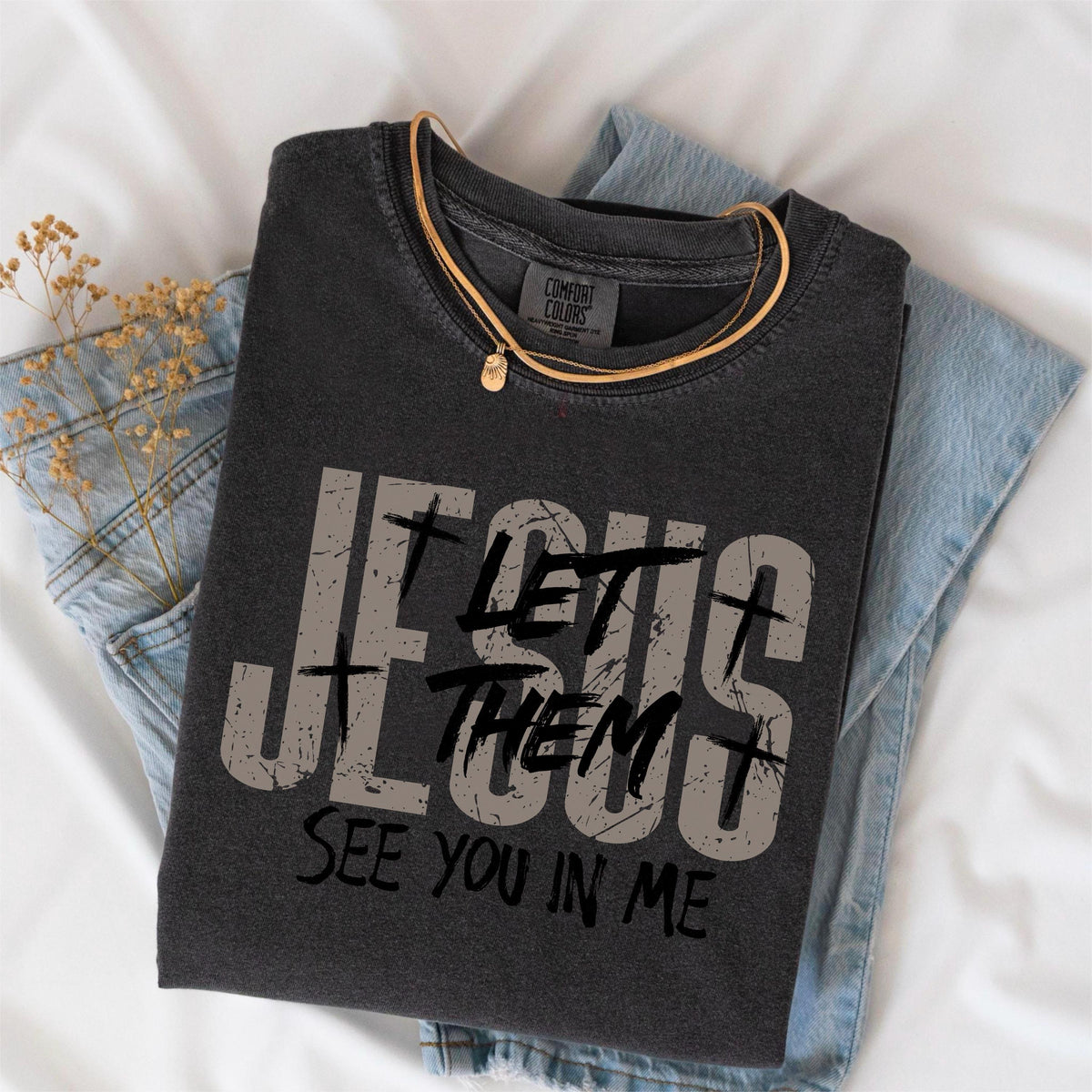 May include: Dark gray t-shirt with the text "JESUS LET THEM SEE YOU IN ME" in distressed gray lettering. A gold necklace with a small pendant rests on the shirt, which is folded on top of a pair of blue jeans.