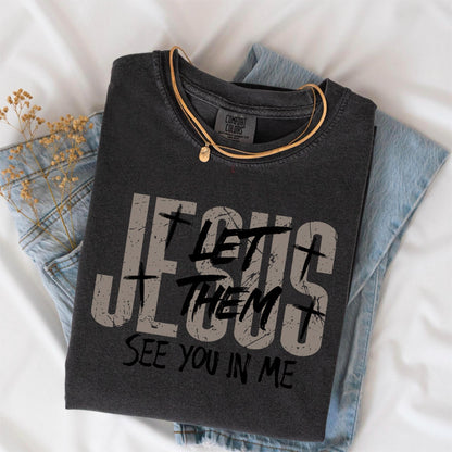 May include: Dark gray t-shirt with the text "JESUS LET THEM SEE YOU IN ME" in distressed gray lettering. A gold necklace with a small pendant rests on the shirt, which is folded on top of a pair of blue jeans.