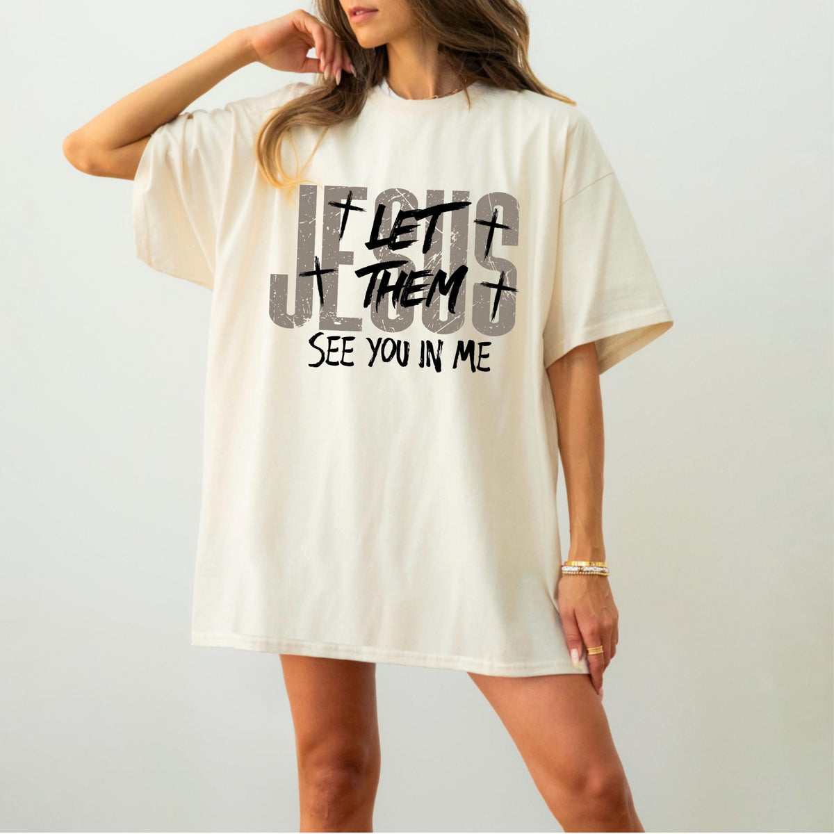 May include: Cream-colored t-shirt with the text "JESUS LET THEM SEE YOU IN ME" in distressed gray and black lettering. The design includes crosses. The shirt has short sleeves and a relaxed fit.