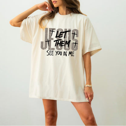 May include: Cream-colored t-shirt with the text "JESUS LET THEM SEE YOU IN ME" in distressed gray and black lettering. The design includes crosses. The shirt has short sleeves and a relaxed fit.