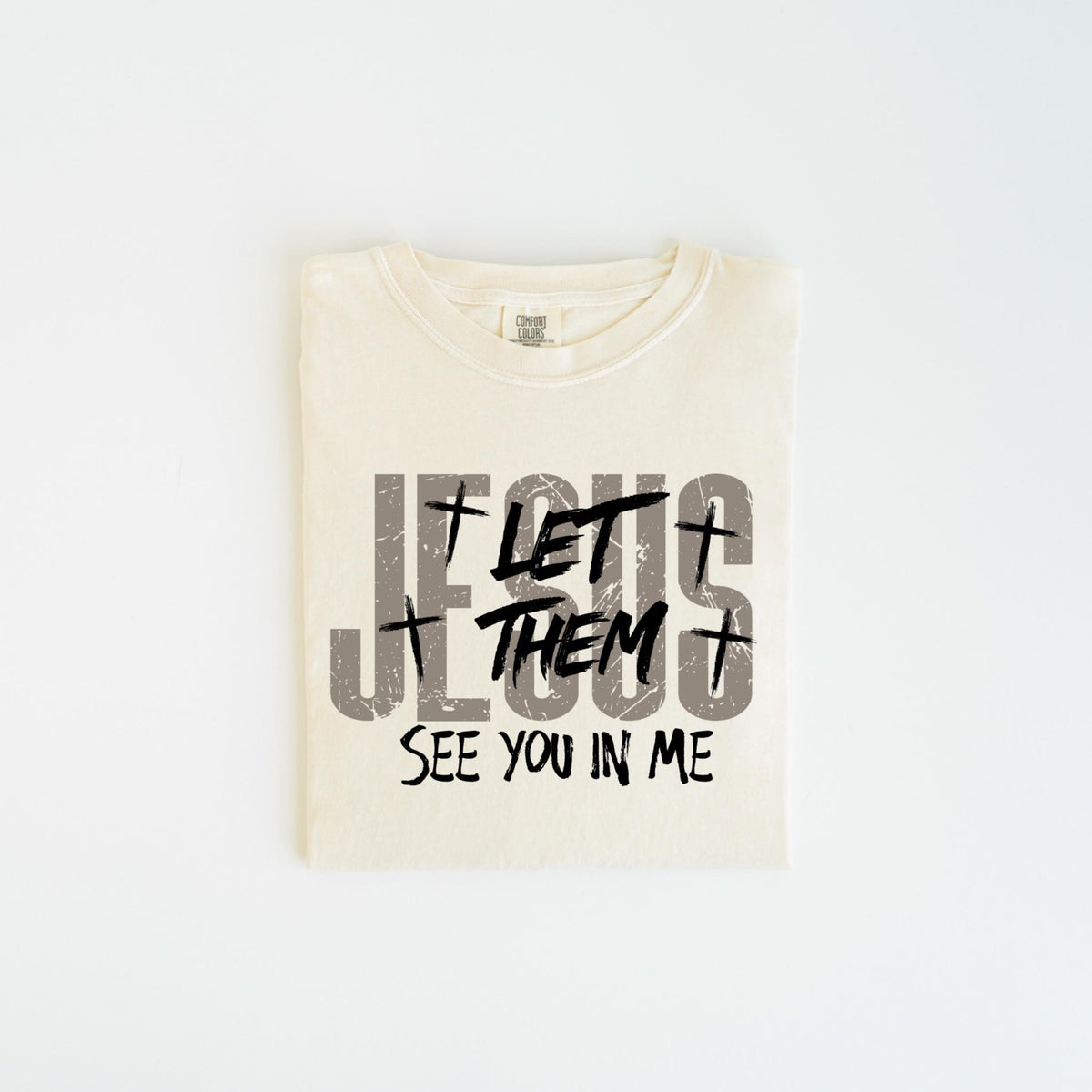 May include: Cream-colored t-shirt with the text "JESUS LET THEM SEE YOU IN ME" in gray and black. The design includes crosses and a distressed font. The shirt is folded and centered on a white background.