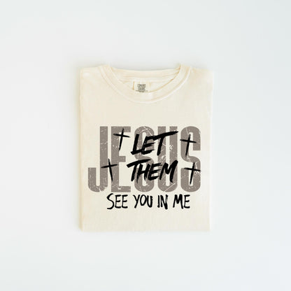 May include: Cream-colored t-shirt with the text "JESUS LET THEM SEE YOU IN ME" in gray and black. The design includes crosses and a distressed font. The shirt is folded and centered on a white background.
