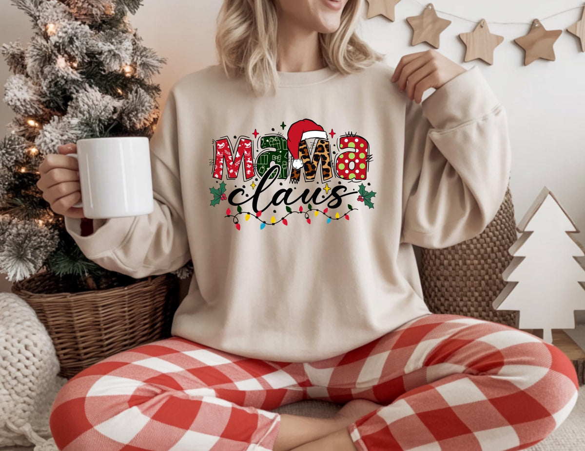 May include: A beige sweatshirt with the text "Mama Claus" in a festive design. The letters are decorated with Christmas patterns, a Santa hat, and string lights. The person is wearing red and white plaid pajama pants and holding a white mug.