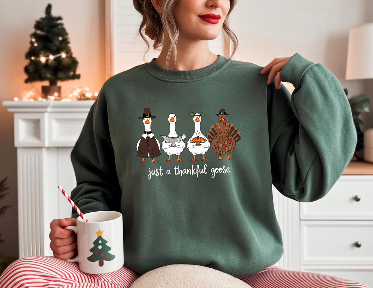 May include: A dark green sweatshirt with a graphic of four geese and a turkey, each wearing festive attire. The text "just a thankful goose" is below the illustration. A white mug with a Christmas tree design is held in the hand.