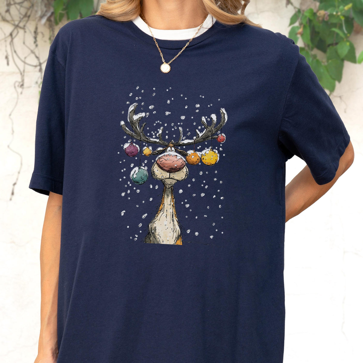 May include: Navy blue t-shirt featuring a whimsical cartoon reindeer with colorful ornaments on its antlers and a red nose. The reindeer is set against a snowy background. The shirt has a white collar.