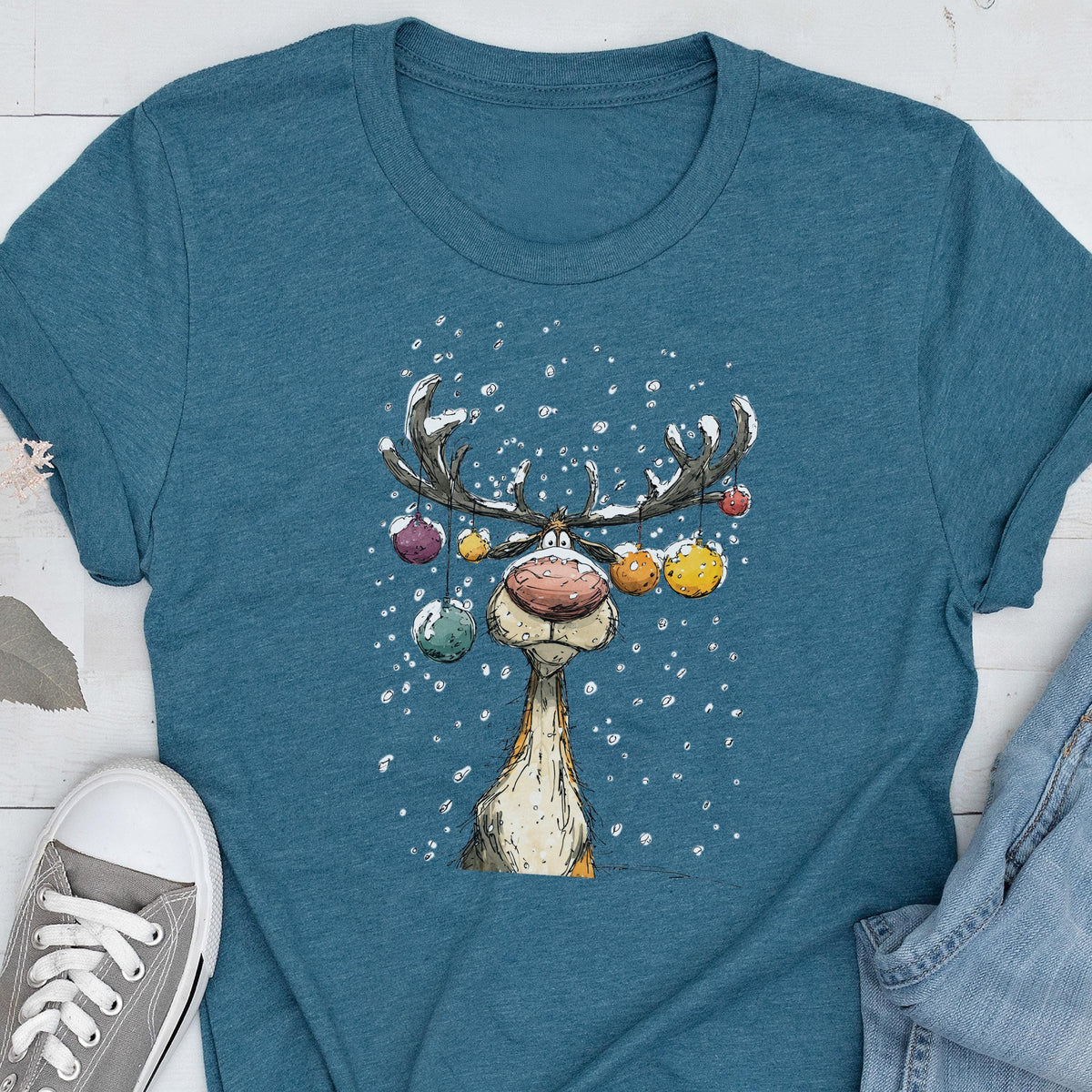 May include: Teal t-shirt featuring a cartoon reindeer with large antlers decorated with colorful ornaments. The reindeer has a large red nose and is surrounded by falling snowflakes. The shirt is a casual, festive design.