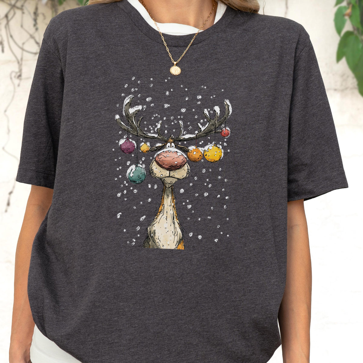 May include: Dark gray t-shirt featuring a whimsical illustration of a reindeer with colorful ornaments hanging from its antlers. The reindeer has a large, textured nose and is set against a snowy background, creating a festive holiday theme.