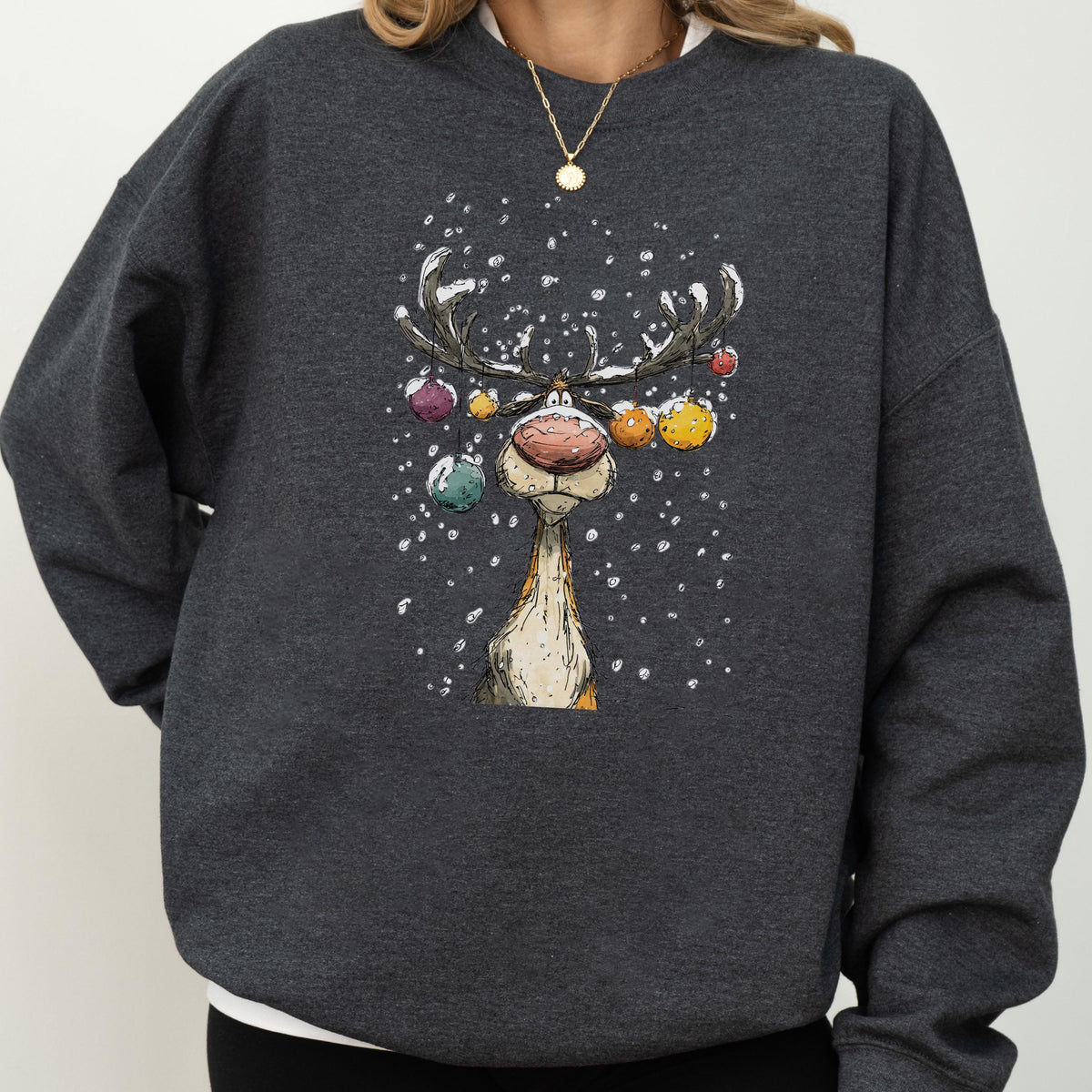 May include: A dark charcoal gray crewneck sweatshirt featuring a graphic illustration of a reindeer with large antlers decorated with colorful hanging ornaments. The reindeer has a prominent nose and is surrounded by white falling snowflakes.