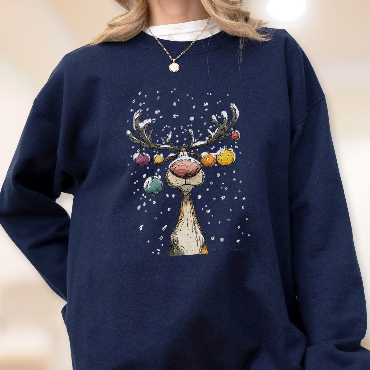 May include: A navy blue crewneck sweatshirt featuring a graphic illustration of a reindeer with a large pink nose. The reindeer has antlers decorated with colorful hanging ornaments, and the design includes white snowflakes against the dark fabric.