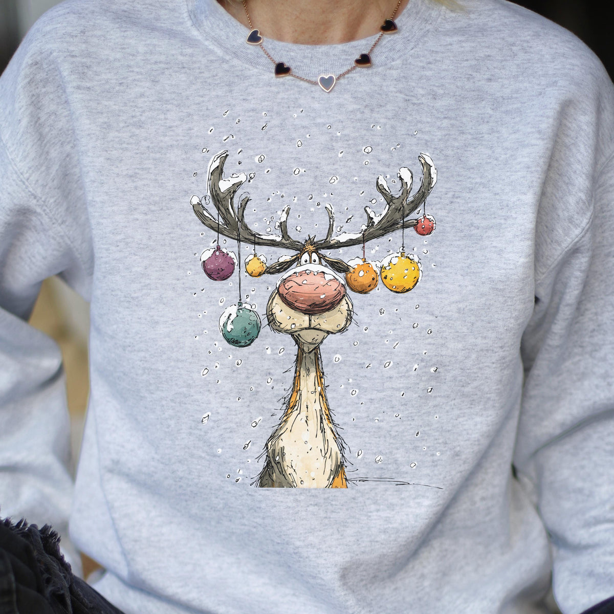 May include: A light gray crewneck sweatshirt featuring a cartoon illustration of a reindeer with large antlers decorated with colorful hanging ornaments. The reindeer has a prominent pink nose and is surrounded by falling white snowflakes.