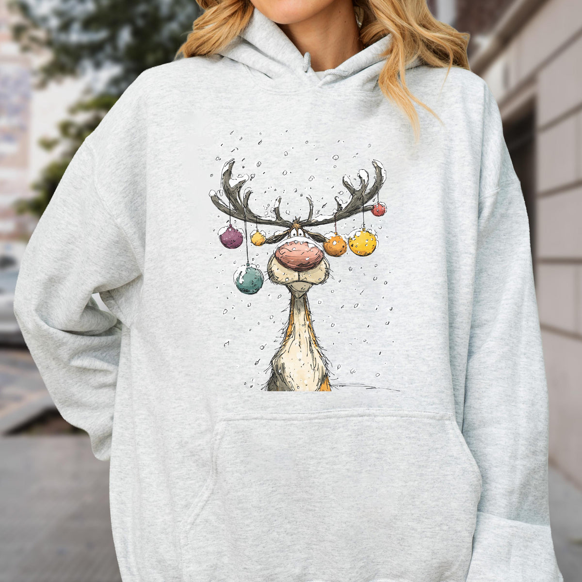 May include: A light gray heather hoodie featuring a graphic illustration of a reindeer with large antlers decorated with colorful hanging ornaments. The reindeer has a prominent pink nose and is surrounded by small white dots resembling falling snow.