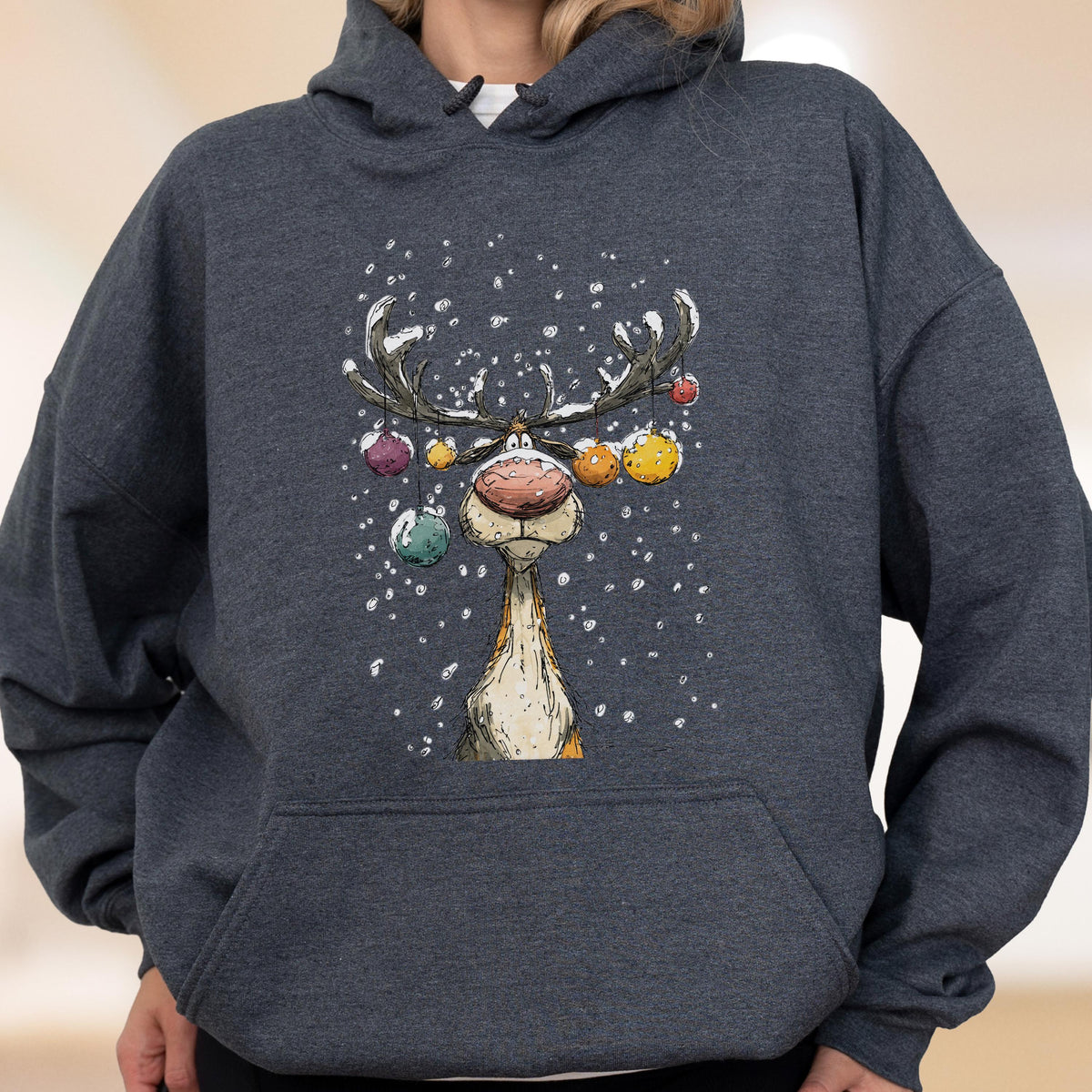 May include: A dark charcoal gray hooded sweatshirt featuring a graphic illustration of a reindeer with a large pink nose. The reindeer has antlers decorated with colorful hanging ornaments and is surrounded by white falling snowflakes.