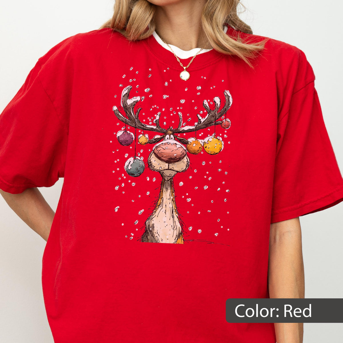 May include: A bright red short-sleeved t-shirt featuring a graphic illustration of a reindeer with large antlers decorated with colorful hanging ornaments. The design includes white specks representing falling snow against the red fabric background.