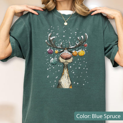 May include: A blue spruce short-sleeve t-shirt featuring a graphic illustration of a reindeer with a large nose and antlers decorated with colorful hanging ornaments, set against a background of falling white snow, with a gold-toned necklace.