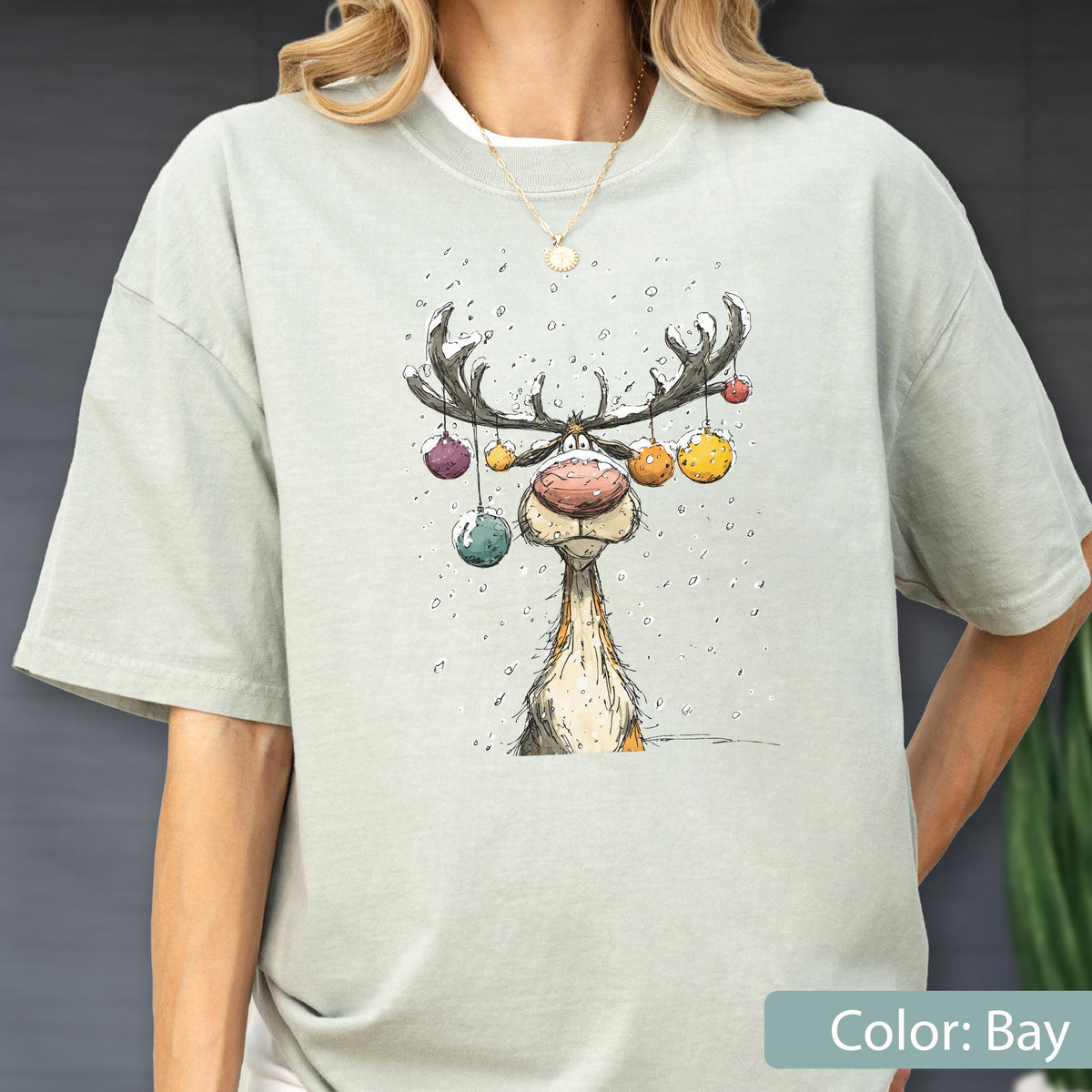 May include: A light sage green short-sleeve t-shirt featuring a whimsical cartoon illustration of a reindeer with large antlers decorated with colorful hanging ornaments, set against a backdrop of falling snow, with a gold chain necklace.