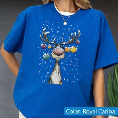 May include: A royal cariba blue t-shirt featuring a graphic illustration of a reindeer with large antlers decorated with colorful hanging ornaments. The reindeer has a prominent pink nose and is surrounded by white falling snowflakes.