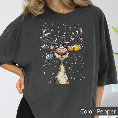 May include: A charcoal gray t-shirt featuring a graphic illustration of a reindeer with a large pink nose and antlers decorated with colorful hanging ornaments. The design includes white specks representing falling snow against the dark fabric.