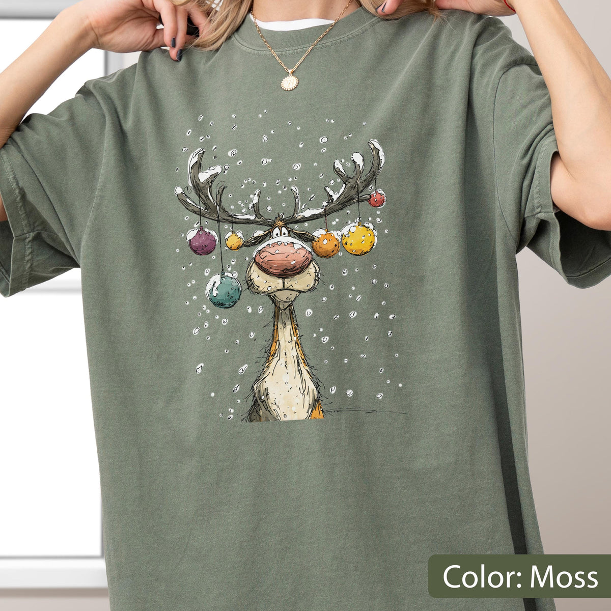 May include: A moss green short-sleeve t-shirt featuring a graphic illustration of a reindeer with large antlers decorated with colorful hanging ornaments. The reindeer has a large nose and is surrounded by white falling snow against the fabric.