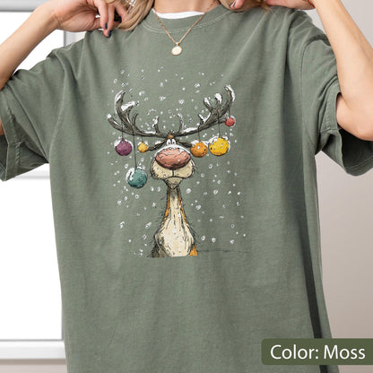 May include: A moss green short-sleeve t-shirt featuring a graphic illustration of a reindeer with large antlers decorated with colorful hanging ornaments. The reindeer has a large nose and is surrounded by white falling snow against the fabric.