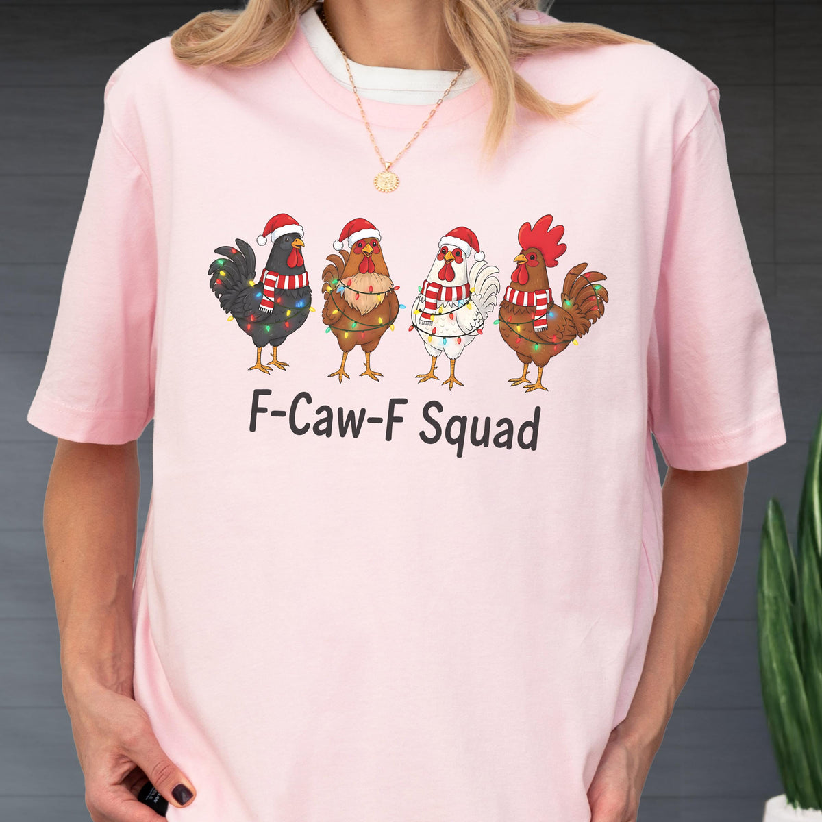 May include: A light pink t-shirt featuring four cartoon roosters wearing Santa hats and Christmas lights. Each rooster has a different color scheme. The text "F-Caw-F Squad" is printed below the roosters.