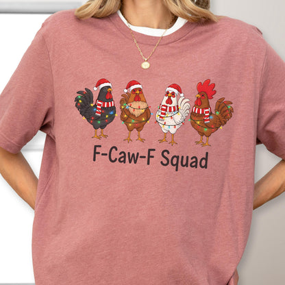 May include: A dusty rose-colored t-shirt featuring a graphic of four cartoon roosters wearing Santa hats and scarves, adorned with Christmas lights. The text "F-Caw-F Squad" is printed below the roosters.