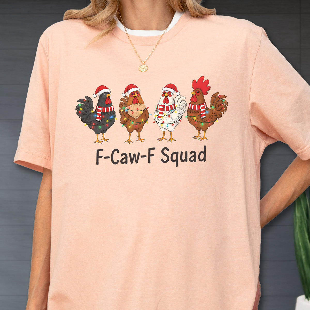 May include: A light pink t-shirt featuring a graphic of four cartoon chickens wearing Santa hats and scarves, adorned with Christmas lights. The text "F-Caw-F Squad" is printed below the chickens.