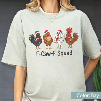 May include: A light green t-shirt featuring four cartoon roosters wearing Santa hats and Christmas lights. The text "F-Caw-F Squad" is printed below the roosters. The shirt is a solid color and has short sleeves.