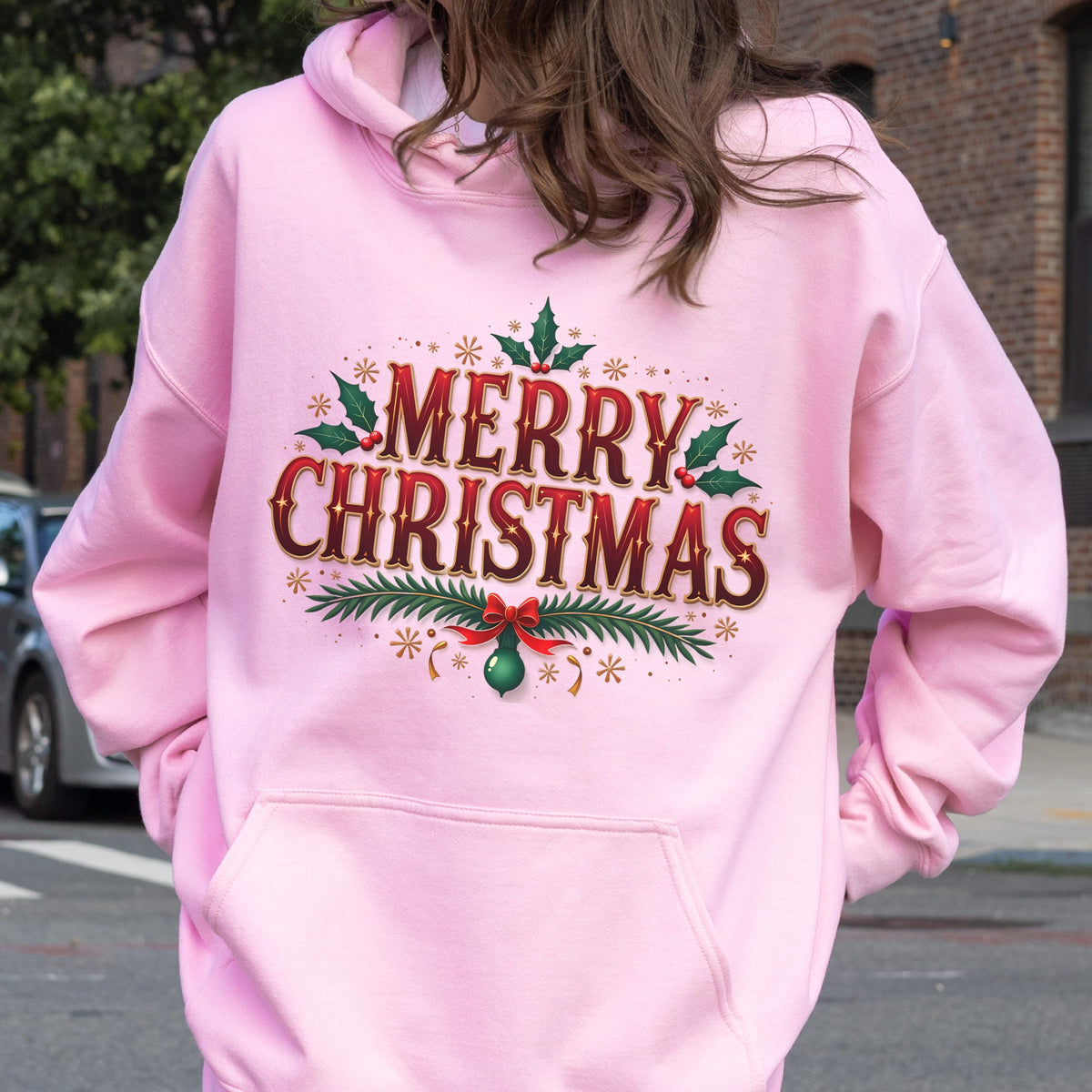 May include: A light pink hoodie with a front pocket and a hood. The back of the hoodie features the words "MERRY CHRISTMAS" in a festive design with red and green accents, holly leaves, and a bow.