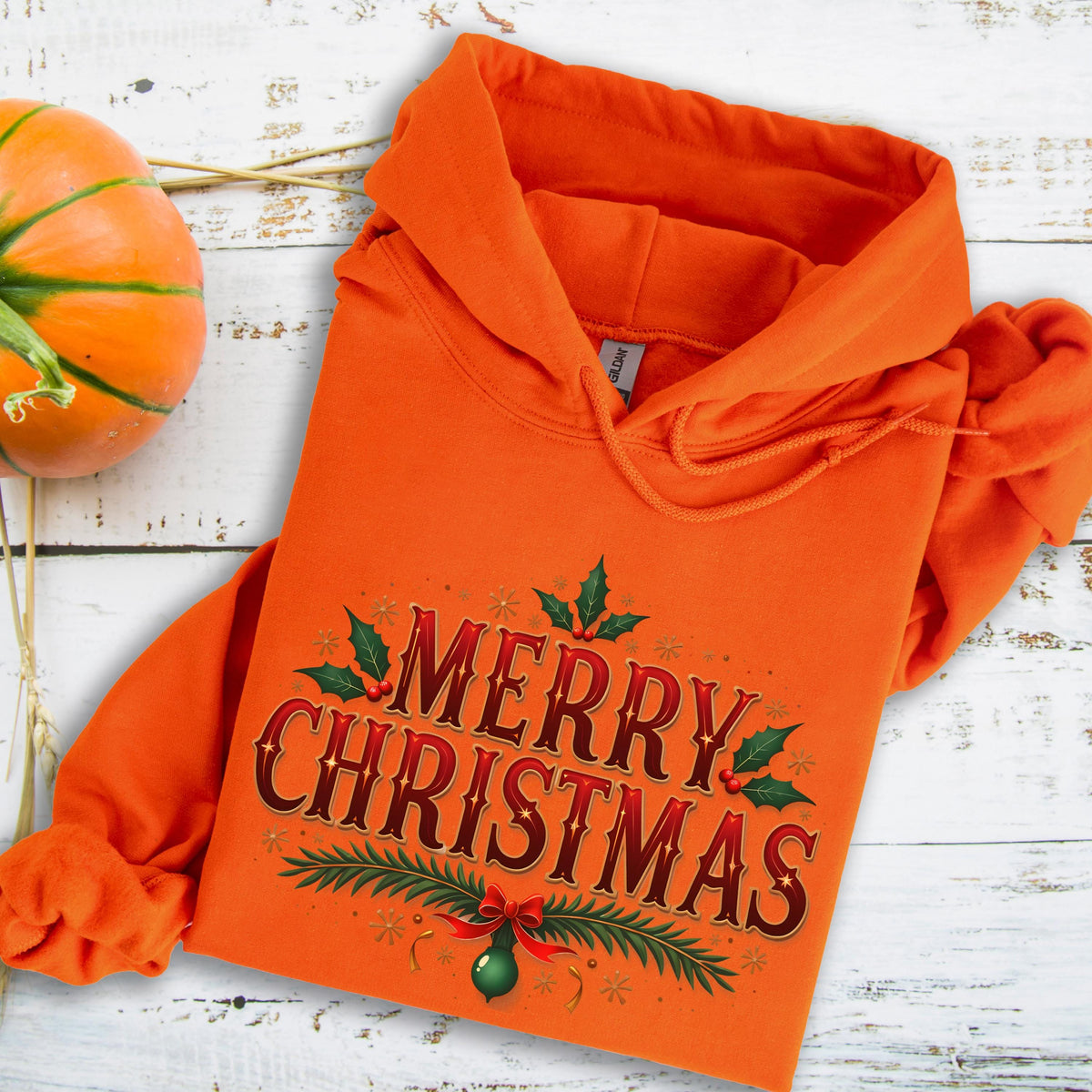 May include: An orange hooded sweatshirt with the words "MERRY CHRISTMAS" in a decorative font. The design includes holly leaves, berries, and a green ornament with a red bow. A pumpkin is visible in the upper left corner.
