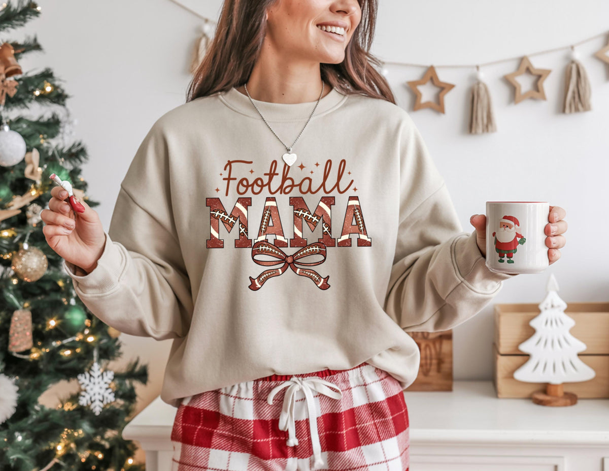 May include: Beige sweatshirt with the text "Football MAMA" in brown and red, featuring football-themed letters and a bow. The person is holding a white mug with a Santa Claus design. The person is wearing red and white plaid pajama pants.