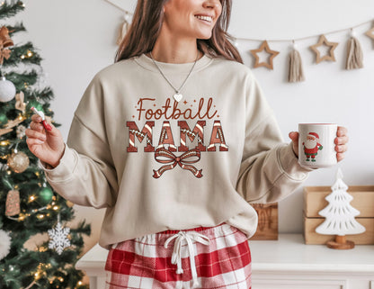 May include: Beige sweatshirt with the text "Football MAMA" in brown and red, featuring football-themed letters and a bow. The person is holding a white mug with a Santa Claus design. The person is wearing red and white plaid pajama pants.