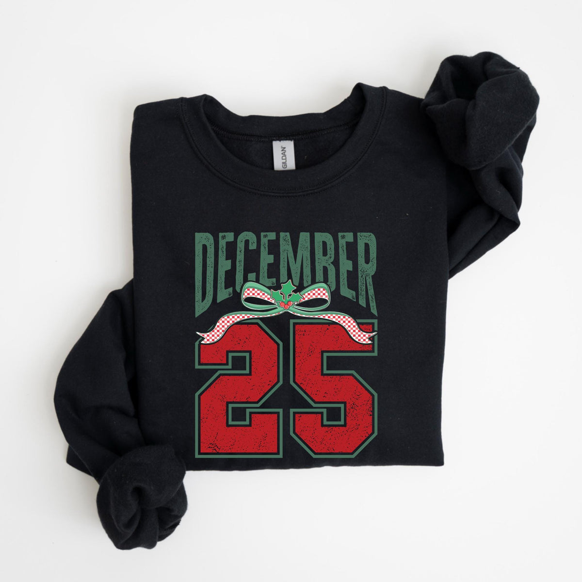 May include: Black sweatshirt with the text "DECEMBER 25" in distressed green and red lettering. A green bow with red and white ribbon accents the text. The sweatshirt is folded on a white surface.