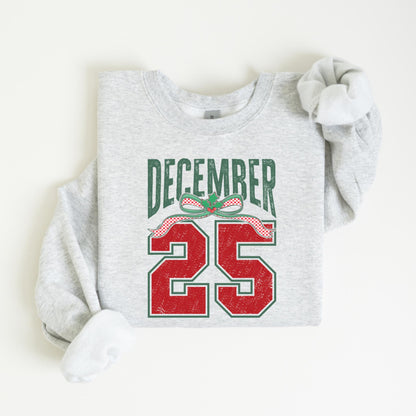 May include: A light gray sweatshirt with the words "DECEMBER 25" printed on the front. The text is in green and red, with a festive bow design. The sweatshirt has long sleeves and a crew neck.