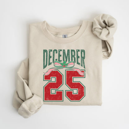 May include: A beige sweatshirt with the text "DECEMBER 25" in green and red. The text is stylized with a bow and ribbon design. The sweatshirt is folded with the sleeves and scrunchie visible.