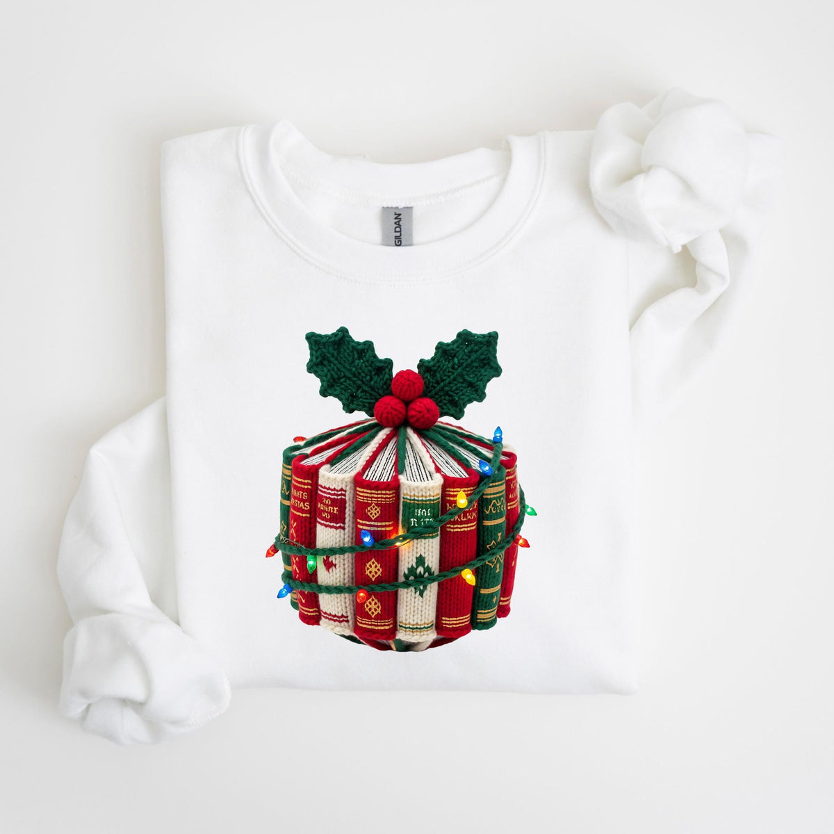 May include: White crew neck sweatshirt featuring a festive design. The design includes a stack of books wrapped with string lights, topped with green holly leaves and red berries. The books are red and green.